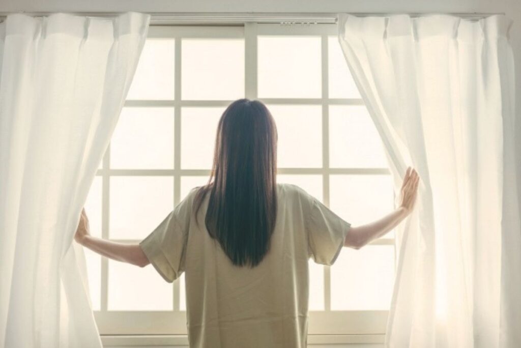 明るい窓辺でカーテンを開ける女性の後ろ姿。自分らしい選択を大切にするイメージ