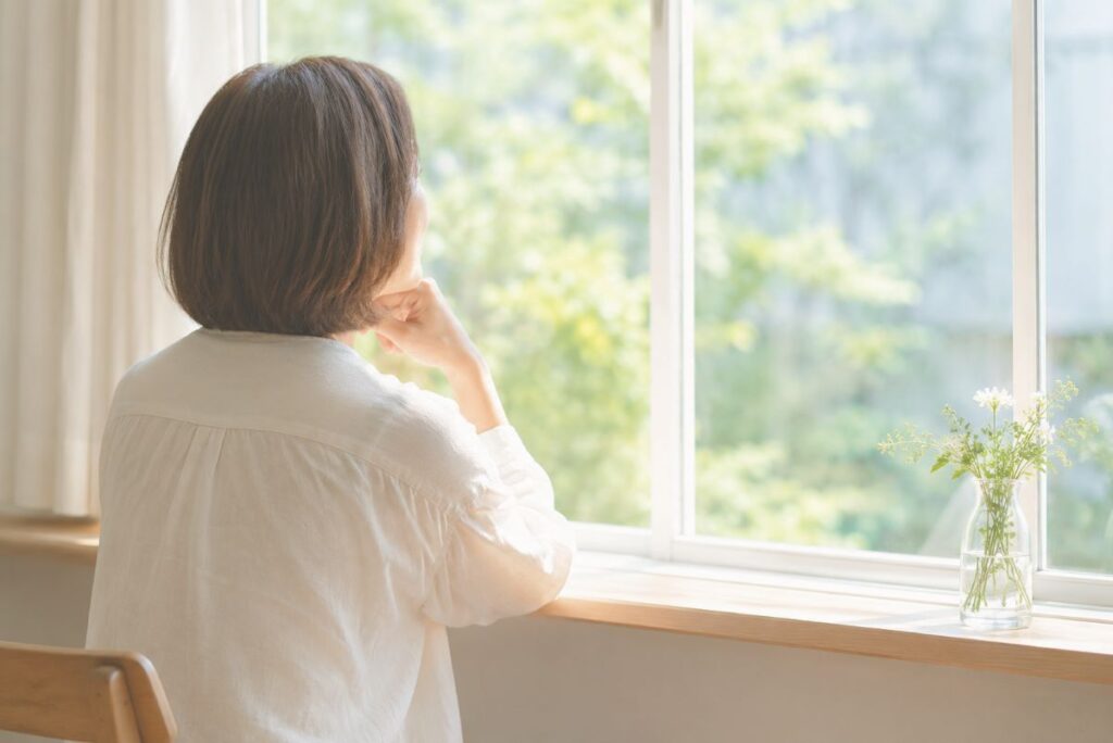明るい窓辺で外を眺める女性。乳がんのことを誰に伝えるか考えるイメージ